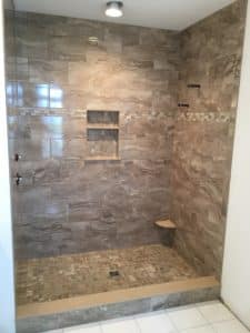 A spacious shower area with textured beige tiles on the walls and a mosaic tile floor. Features include a built-in shelf, a wooden corner seat, and sleek black fixtures. A small horizontal window near the top allows for natural light.