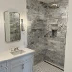 A luxurious bathroom featuring a vanity with a light blue cabinet and marble countertop, an elegant framed mirror, and a spacious walk-in shower with tile walls and floor in various patterns. The shower includes a built-in shelf and a large rain shower head.