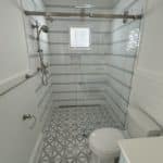 A modern bathroom featuring a large glass-enclosed shower with light gray striped tile walls, a geometric patterned floor, and a rain shower head.