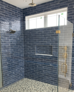 A modern shower with dark blue subway tiles and a pebble tile floor. The design includes a ceiling-mounted rainfall showerhead, a wall-mounted handheld shower, and gold fixtures, complemented by a large glass panel and a window letting in natural light.